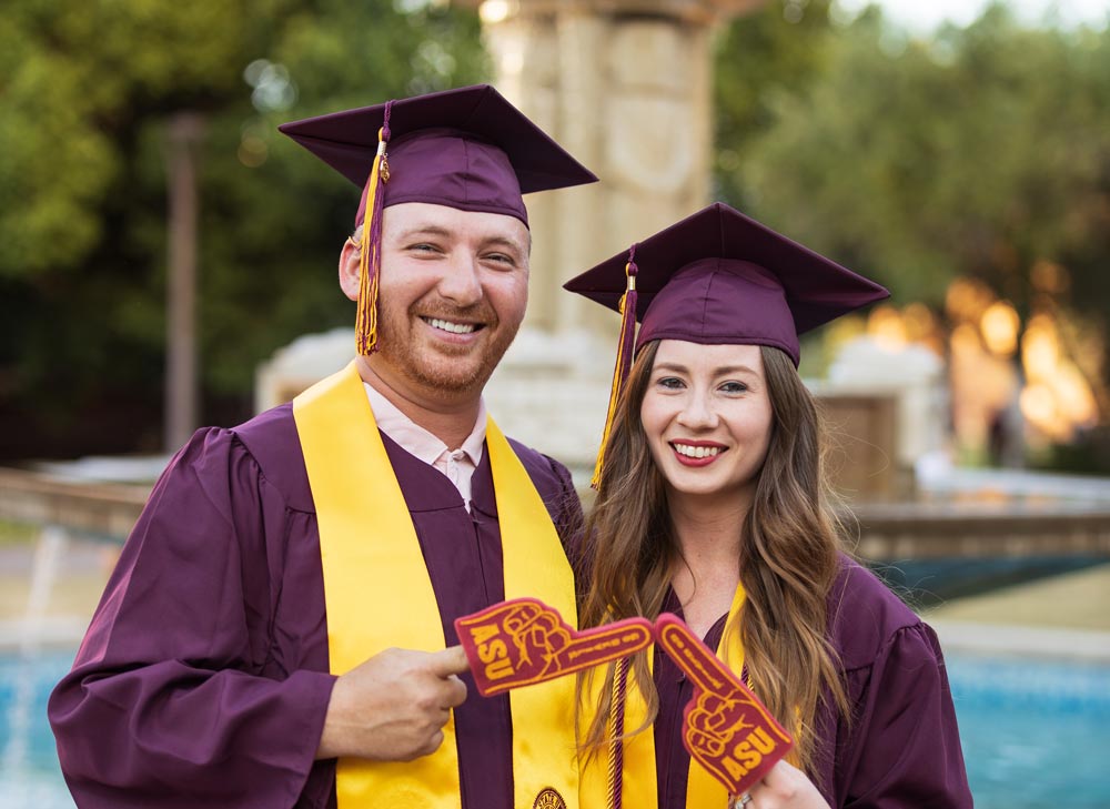 Two ASU Online students dressed in graduation caps and gowns tap together small ASU foam fingers.