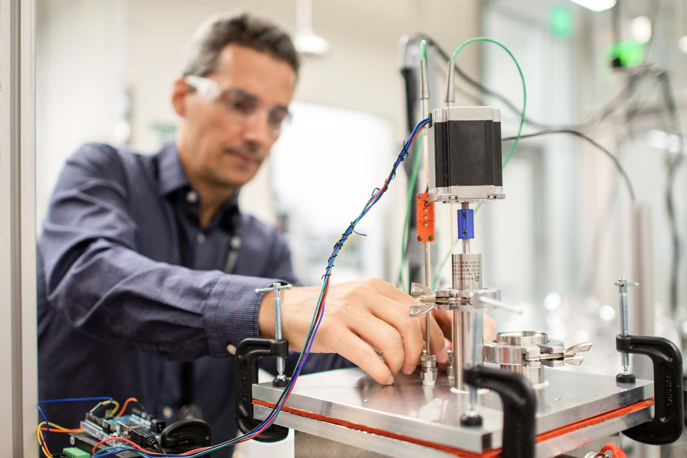 A man with glasses and a blue collared shirt carefully inspects and adjusts wires on a complex machine.