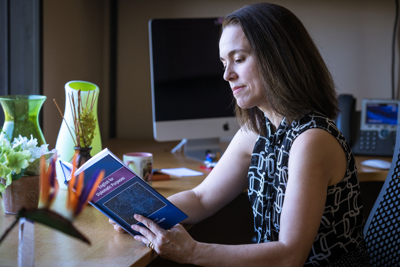 Sitting at her desk surrounded by plants, a woman reads a book.