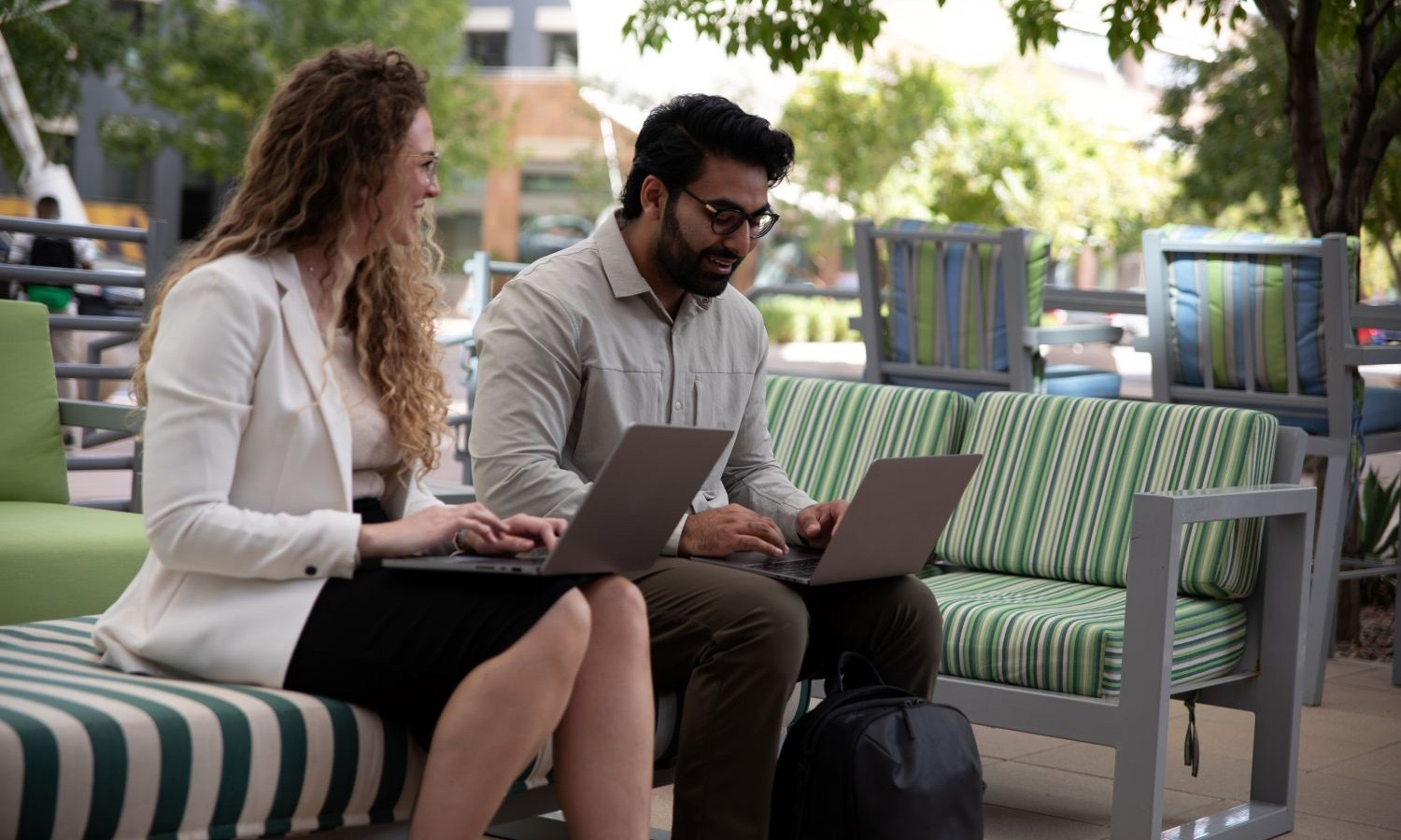 Two students in a management of technology degree program sit outdoors on striped benches and work on laptops.